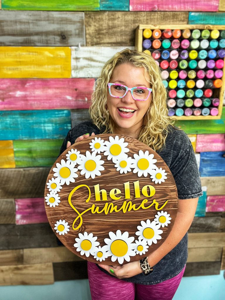 woman holding a door hanger that says "hello summer" with daisies around the edge